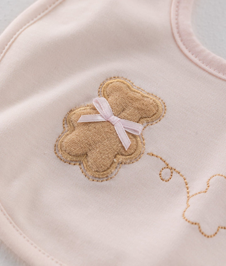 Pink cotton baby bib close-up with tan teddy bear patch, pink ribbon bow, and gold embroidery detail