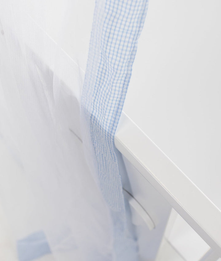 Extreme close-up of blue and white gingham ribbon binding attached to sheer white mesh fabric against white painted wood
